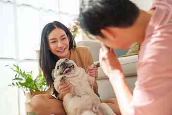 A woman is sitting on a couch with a dog, and a man is standing beside her holding a remote control.